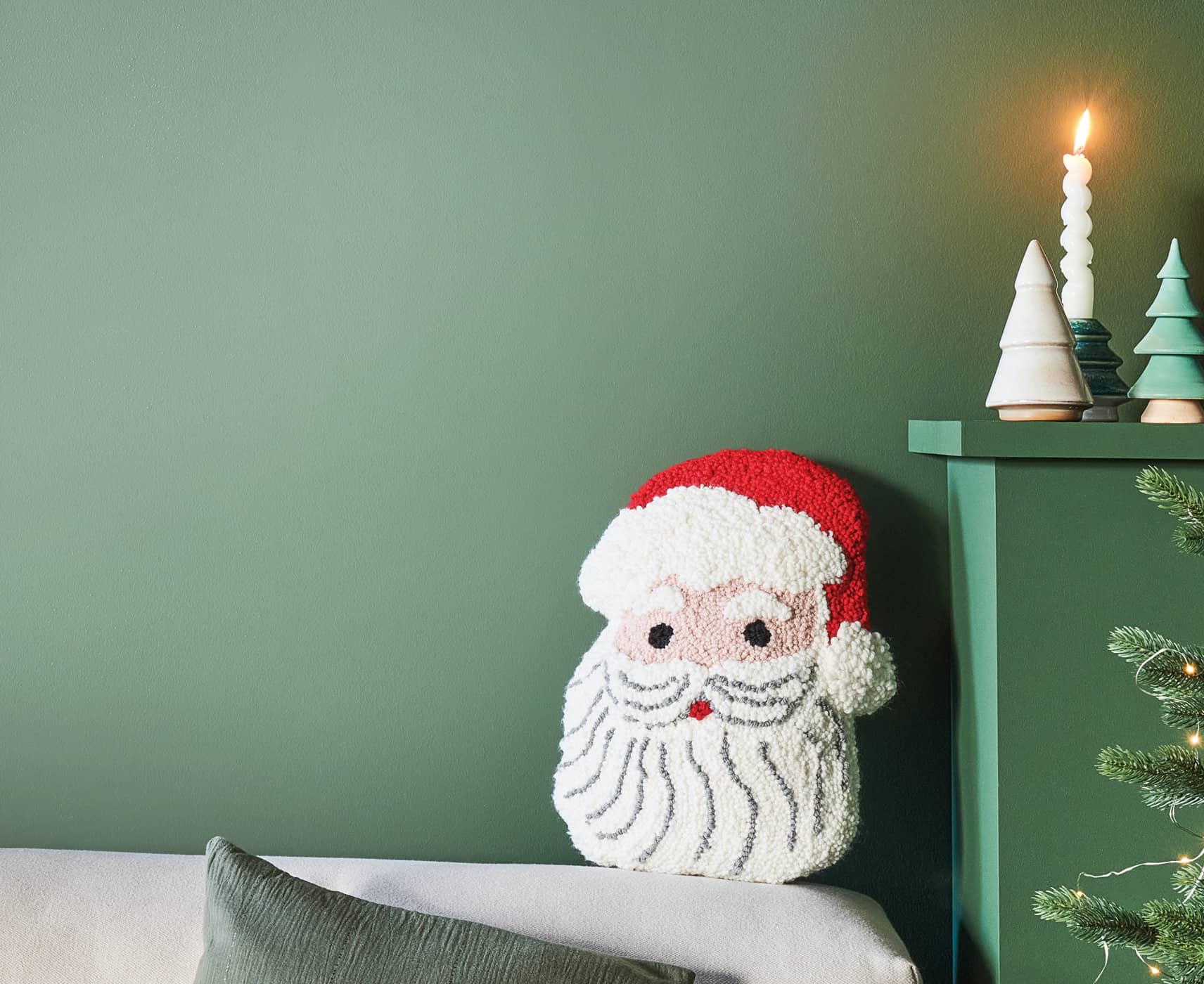Punch-Needle-Kissen in Form eines Weihnachtsmanns auf einem Sofa vor grüner Wand neben einem festlich dekorierten Weihnachtsbaum.