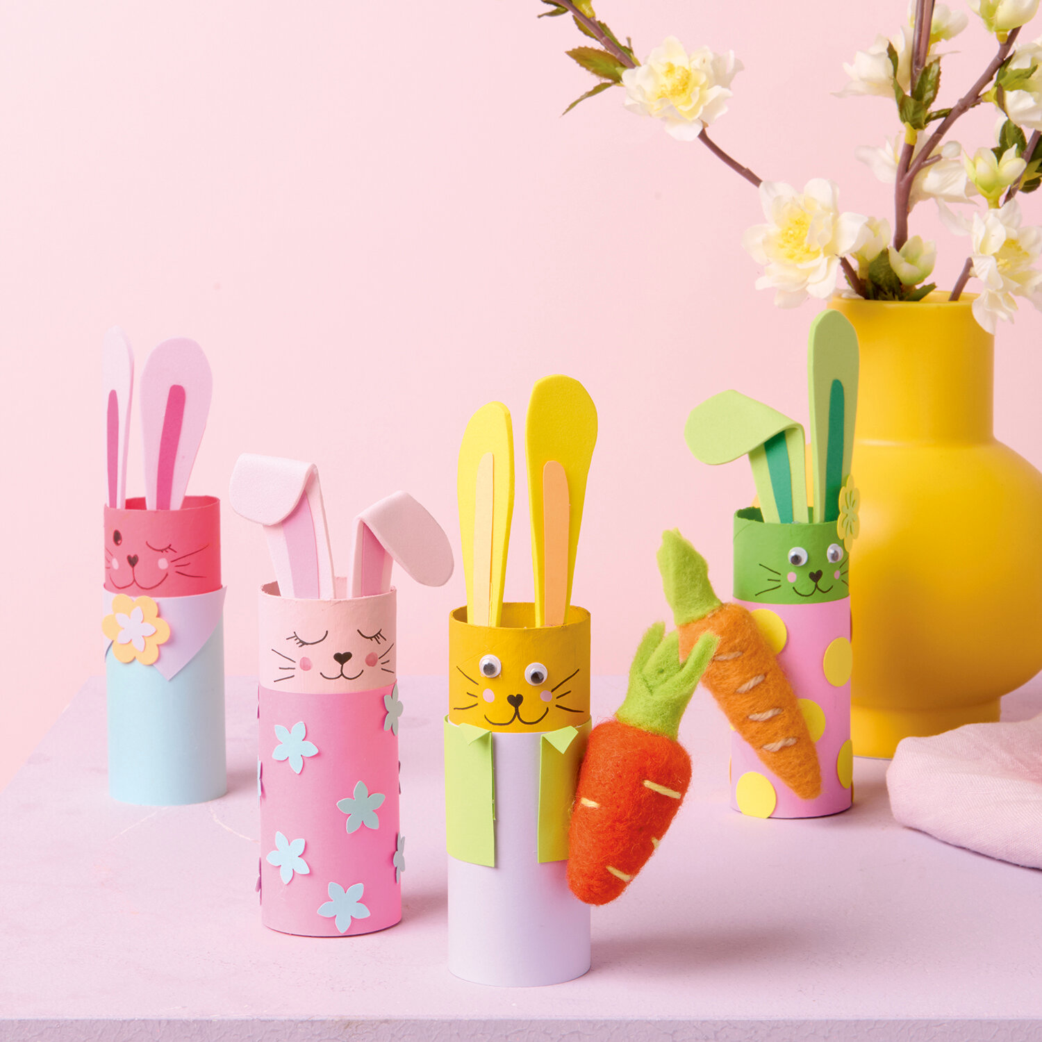 Bastelanleitung Osterhasen aus Klopapierrollen: Bunte Hasenfiguren aus Papierrollen mit Blumen und Karotten auf rosa Hintergrund.