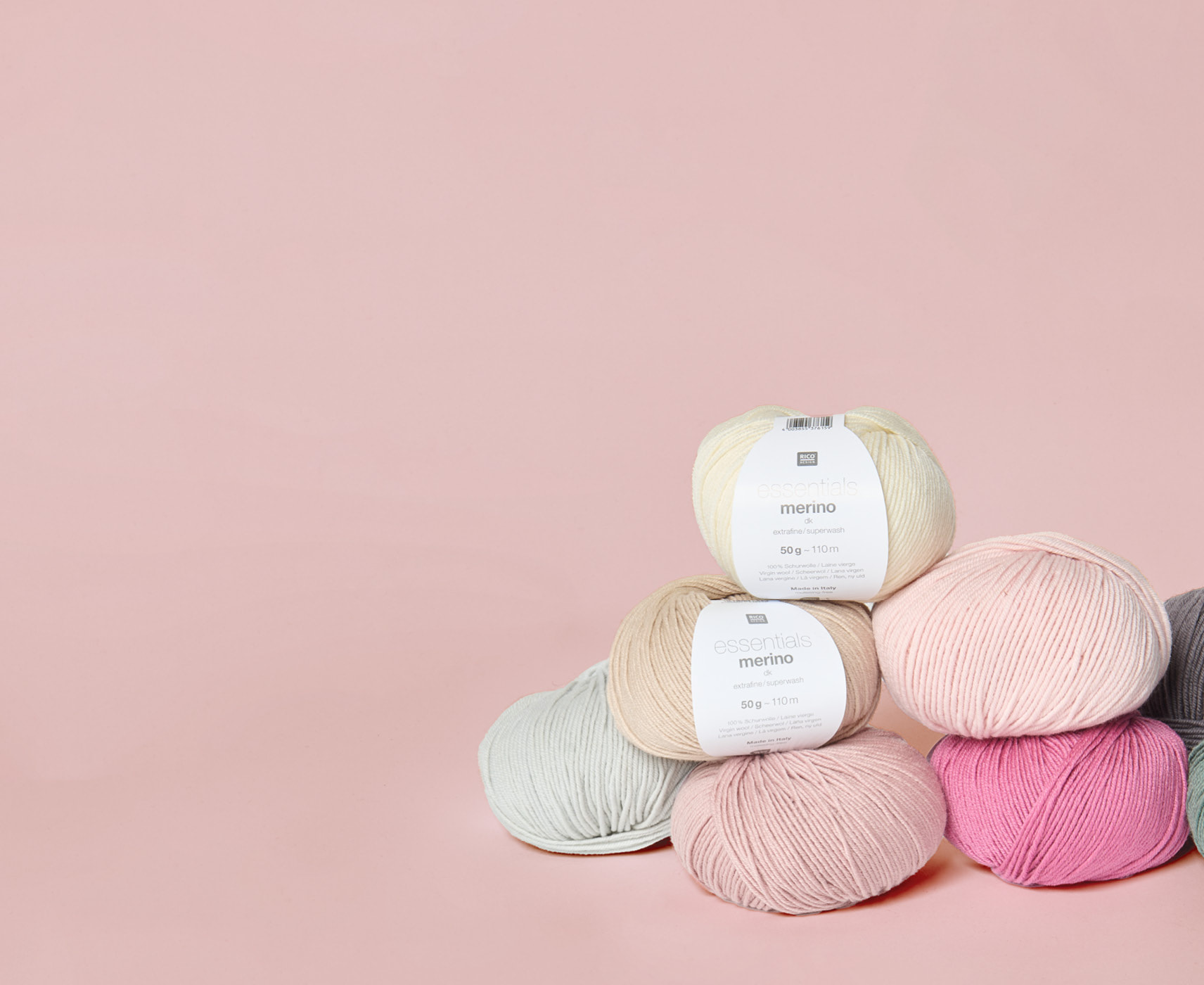 Mehrere Knäuel von Essentials Merino dk in verschiedenen Farben liegen gestapelt vor rosa Hintergrund. 