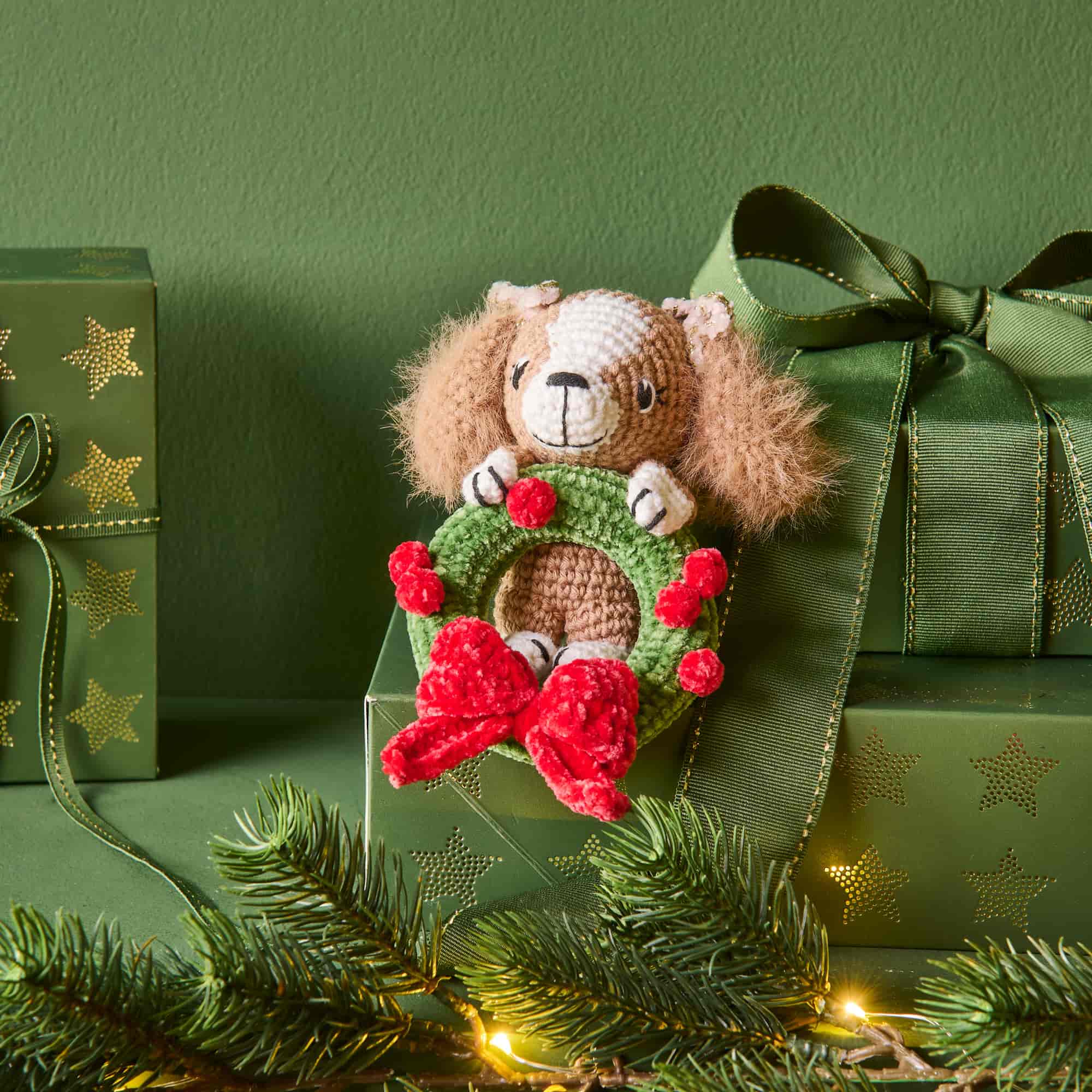 Gehäkelte Figur in Form eines Hundes mit Weihnachtskranz und roter Schleife sitzt vor weihnachtlich dekorierten Geschenken und Tannenzweigen.