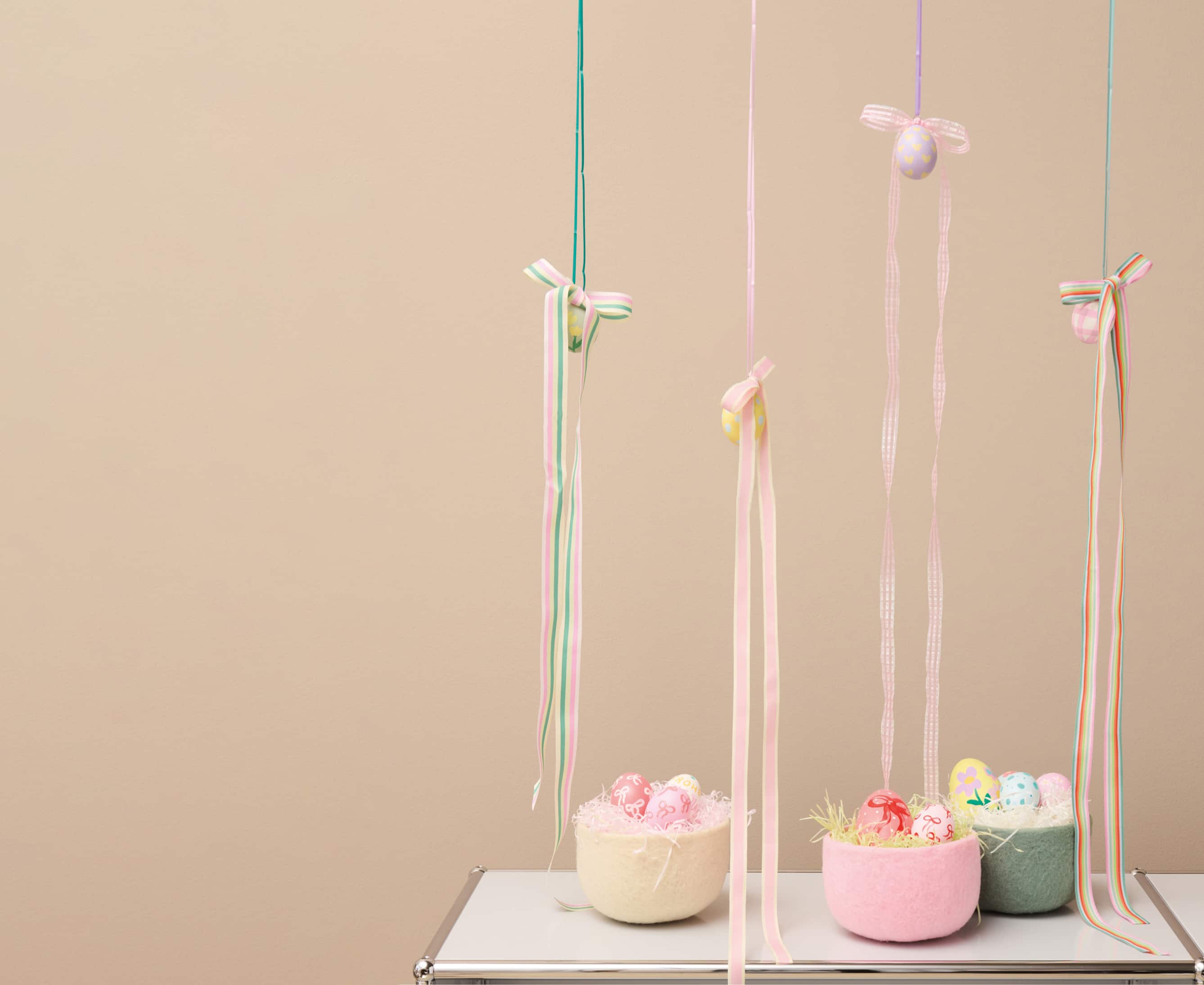 Bemalte Eier sind dekorativ an Geschenkbändern vor einer Wand aufgehangen. Auf einem Regal stehen gefüllte Osterkörbe aus Filz.