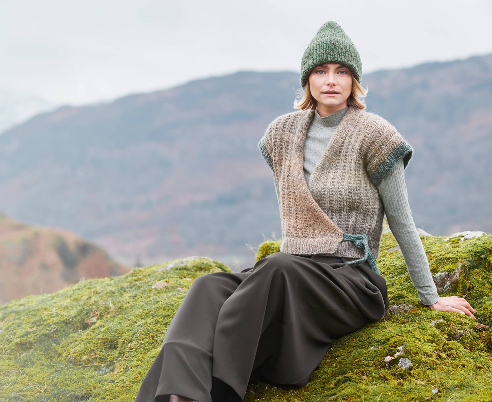 Eine Frau trägt ein herbstliches Outfit mit gestrickter Weste und Mütze und sitzt vor einer Landschaft mit Bergen. 
