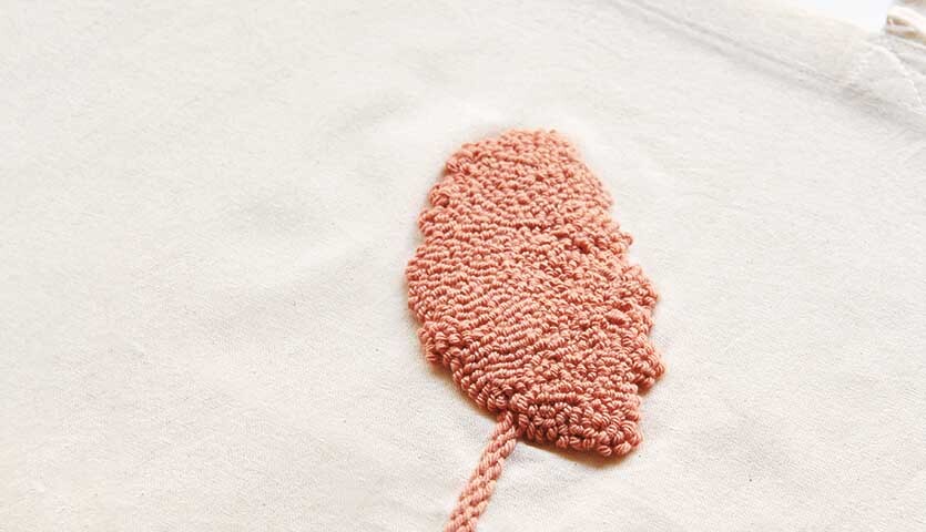 Anleitung Jutebeutel mit der Punch Needle gestalten Anleitung Jutebeutel mit der Punch Needle gestalten: Nahaufnahme eines beigen Stoffes mit rosa Stickerei in Blattform.