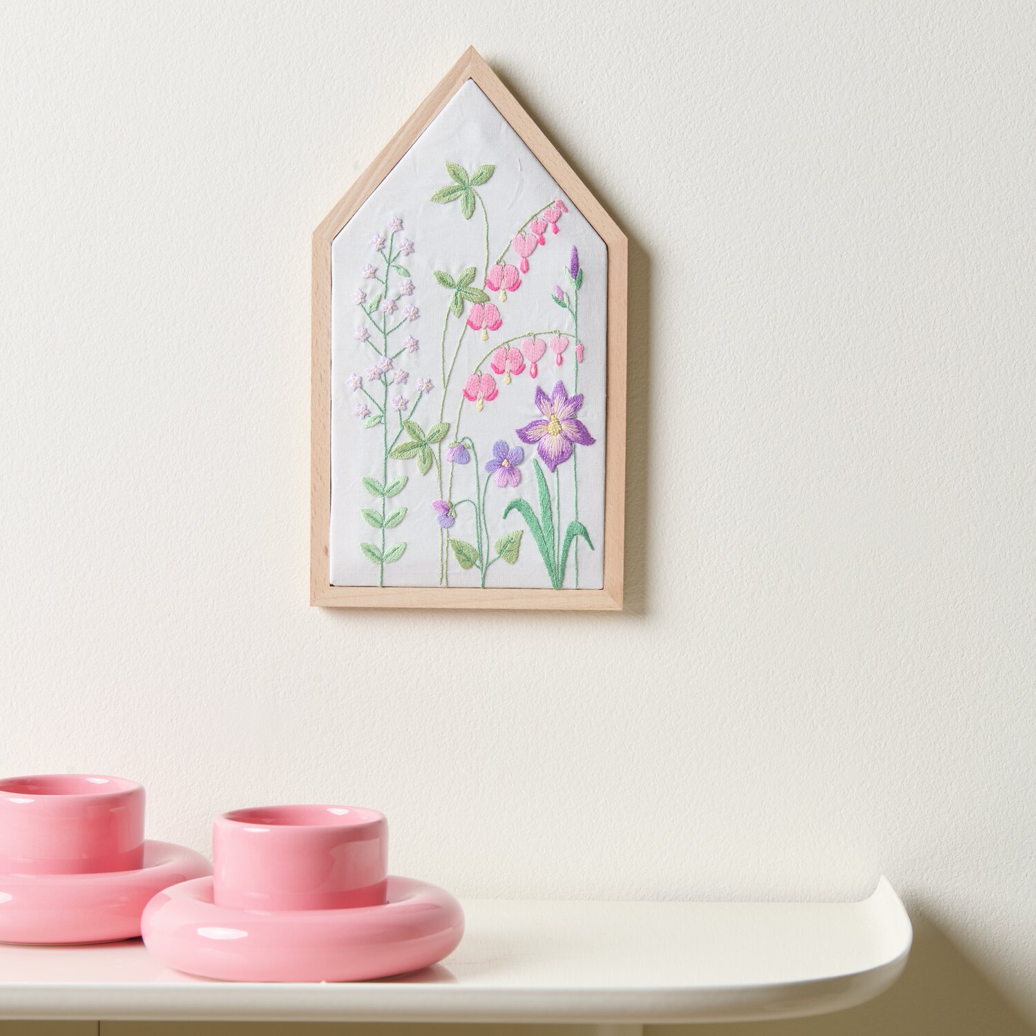 Stickpackung Vorgezeichnet Frühlingshafte Blumenwiese zeigt bunte Blumen in Holzrahmen. Daneben stehen zwei rosa Kerzenhalter auf einem weißen Regal.