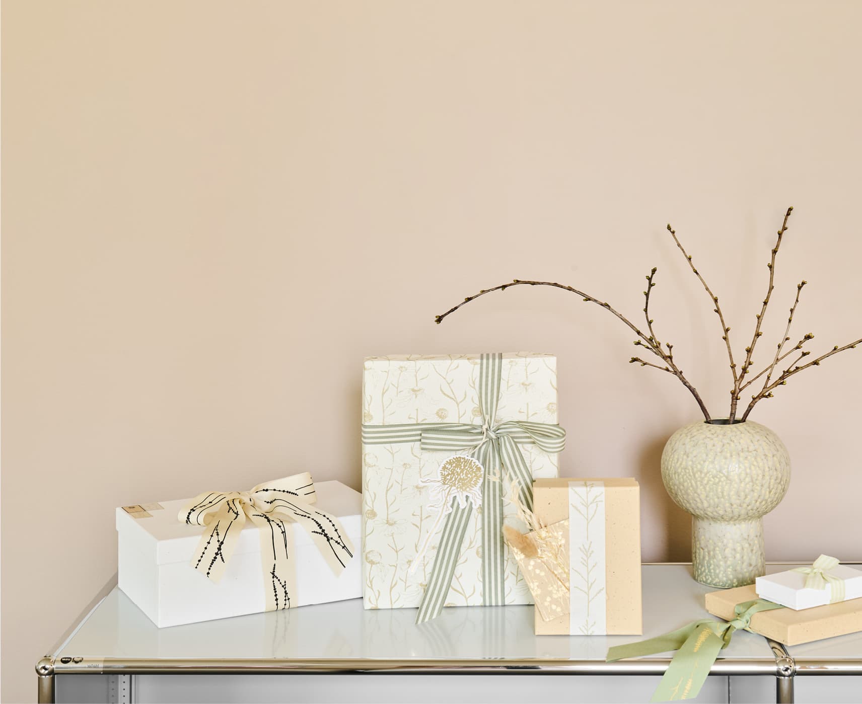 Verpackte Geschenke in Naturtönen und mit Details von Gräsermotiven stehen dekorativ auf einem weißen Regal. 