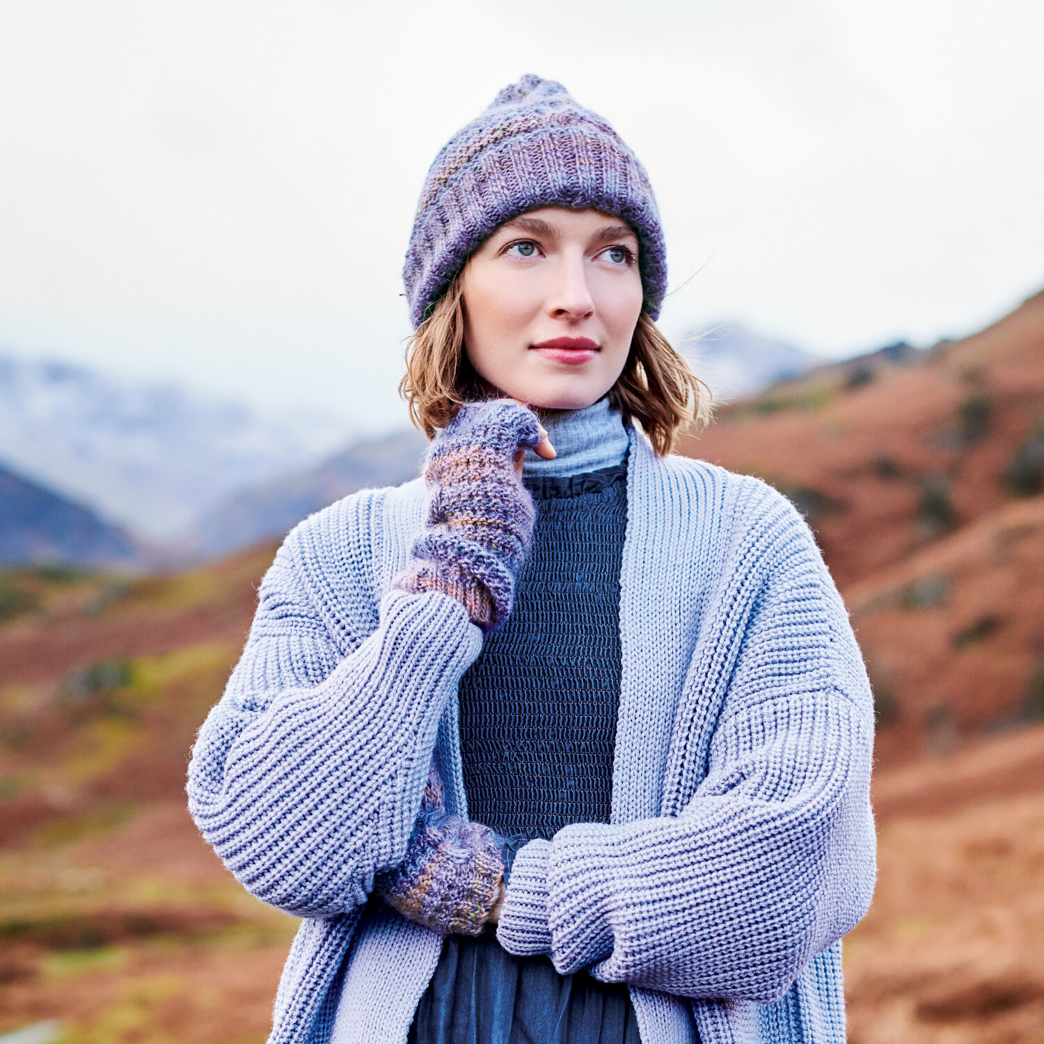 Strickanleitung Mütze und Stulpen aus Creative Alpaca Melange aran zeigt Frau im herbstlichen Bergpanorama mit gestrickter Mütze, Stulpen und Cardigan.