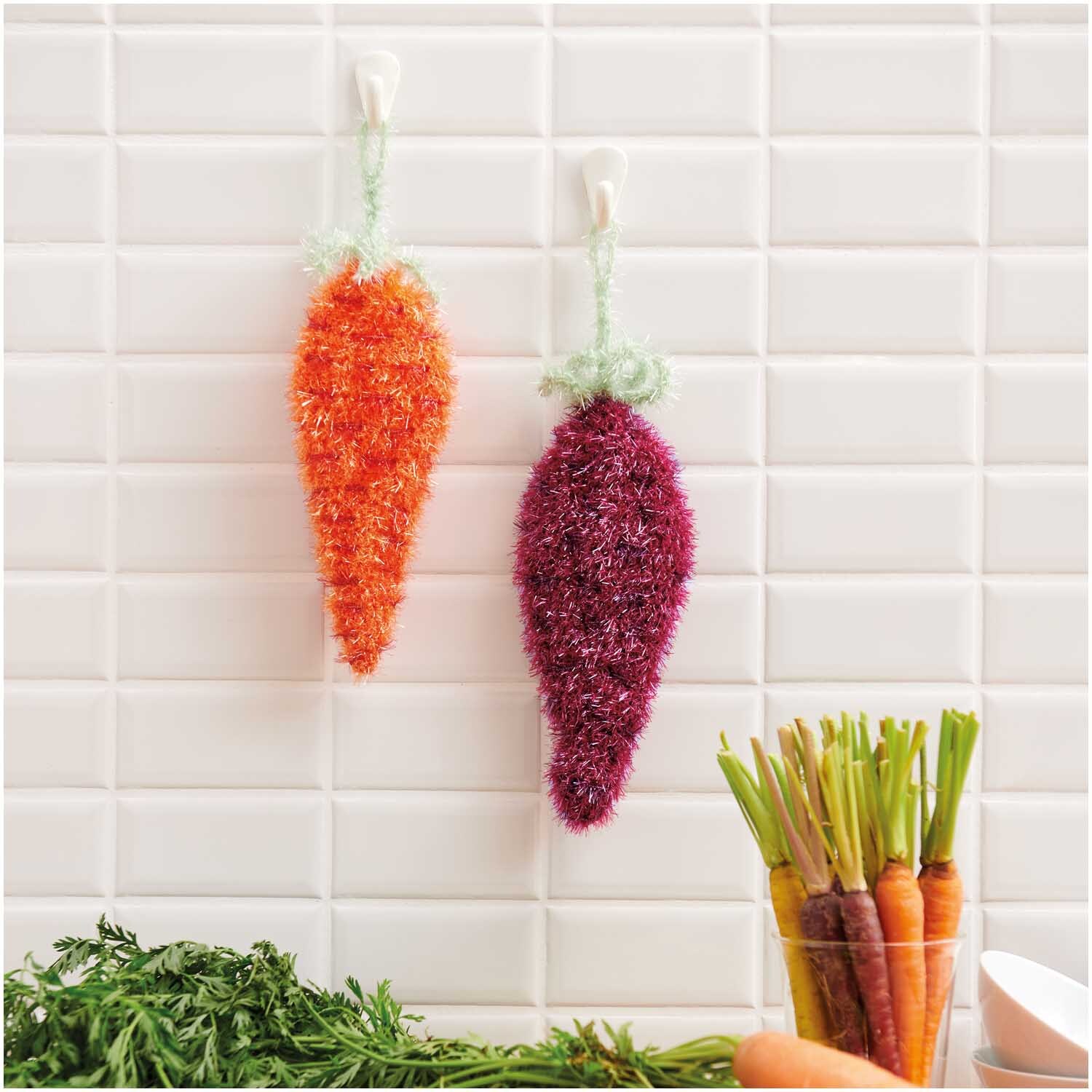 Creative Bubble: Zwei gehäkelte Karotten in Orange und Lila hängen an weißen Fliesen.
