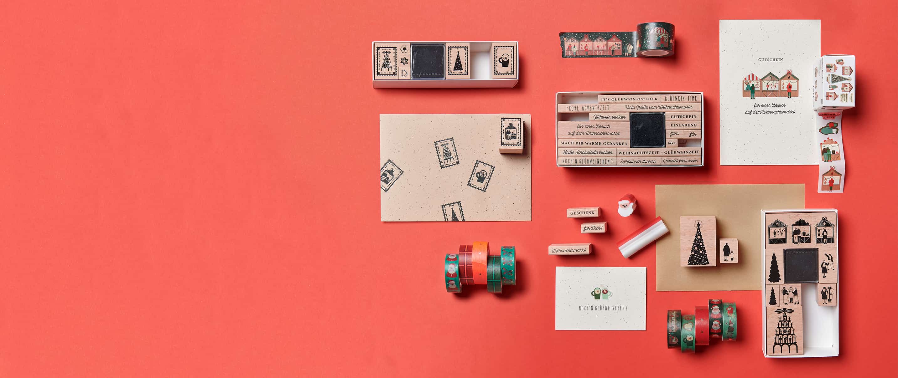 Diverse Stempel mit weihnachtlichen Motiven liegen zusammen mit Weihnachtskarten und Washi Tapes dekoriert auf rotem Hintergrund. 