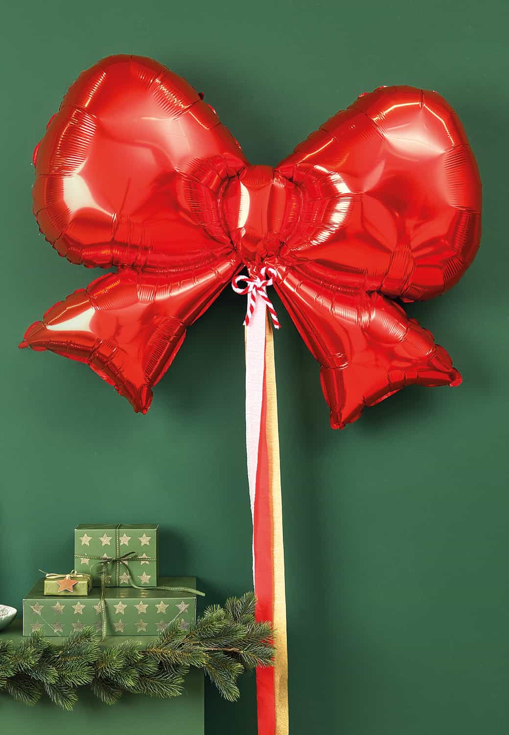 Ein Folienballon in Form einer roten Schleife ist dekorativ vor einer dunkelgrünen Wand platziert. 