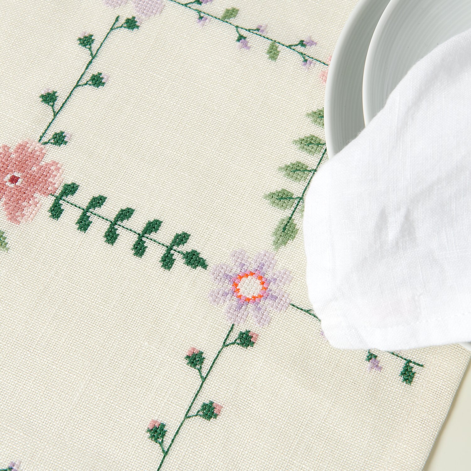 Stickpackung Gezählter Kreuzstich Tischläufer Wiesenblümchen mit floralen Mustern in Grün und Rosa, dekoriert auf einem Tisch mit weißem Geschirr.