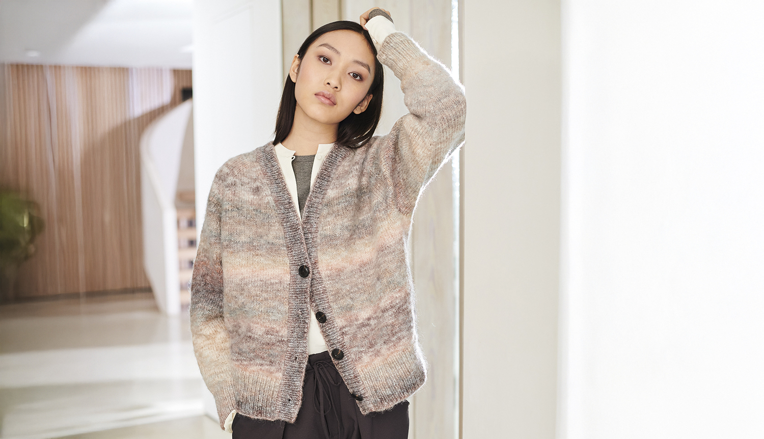 Strickanleitung Jacke aus Creative Painted Power: Frau trägt gestrickte Jacke in sanften Pastelltönen, stehend in modernem Innenraum.