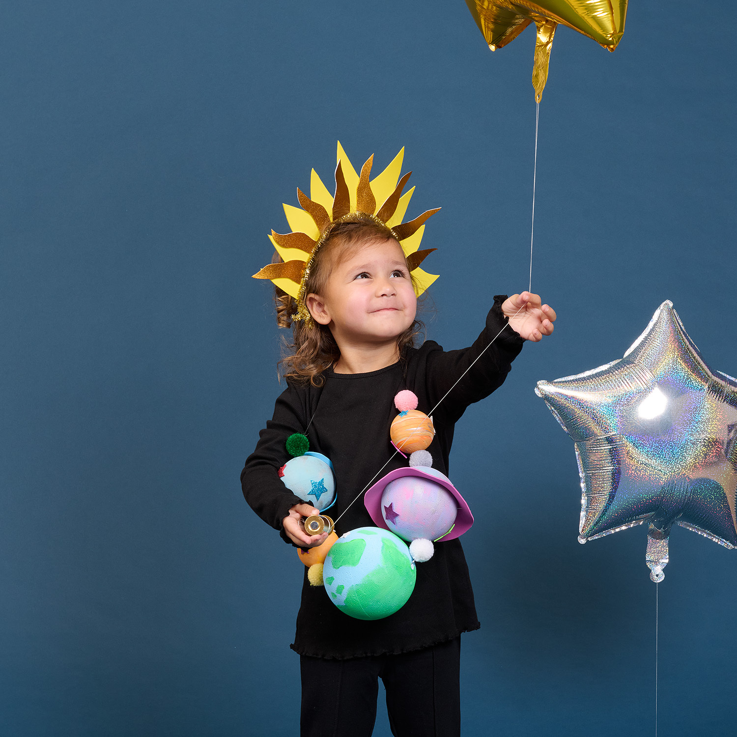Bastelanleitung Planeten-Kostüm: Kind in schwarzem Outfit mit buntem Planeten-Schmuck und goldener Sonnenkrone.