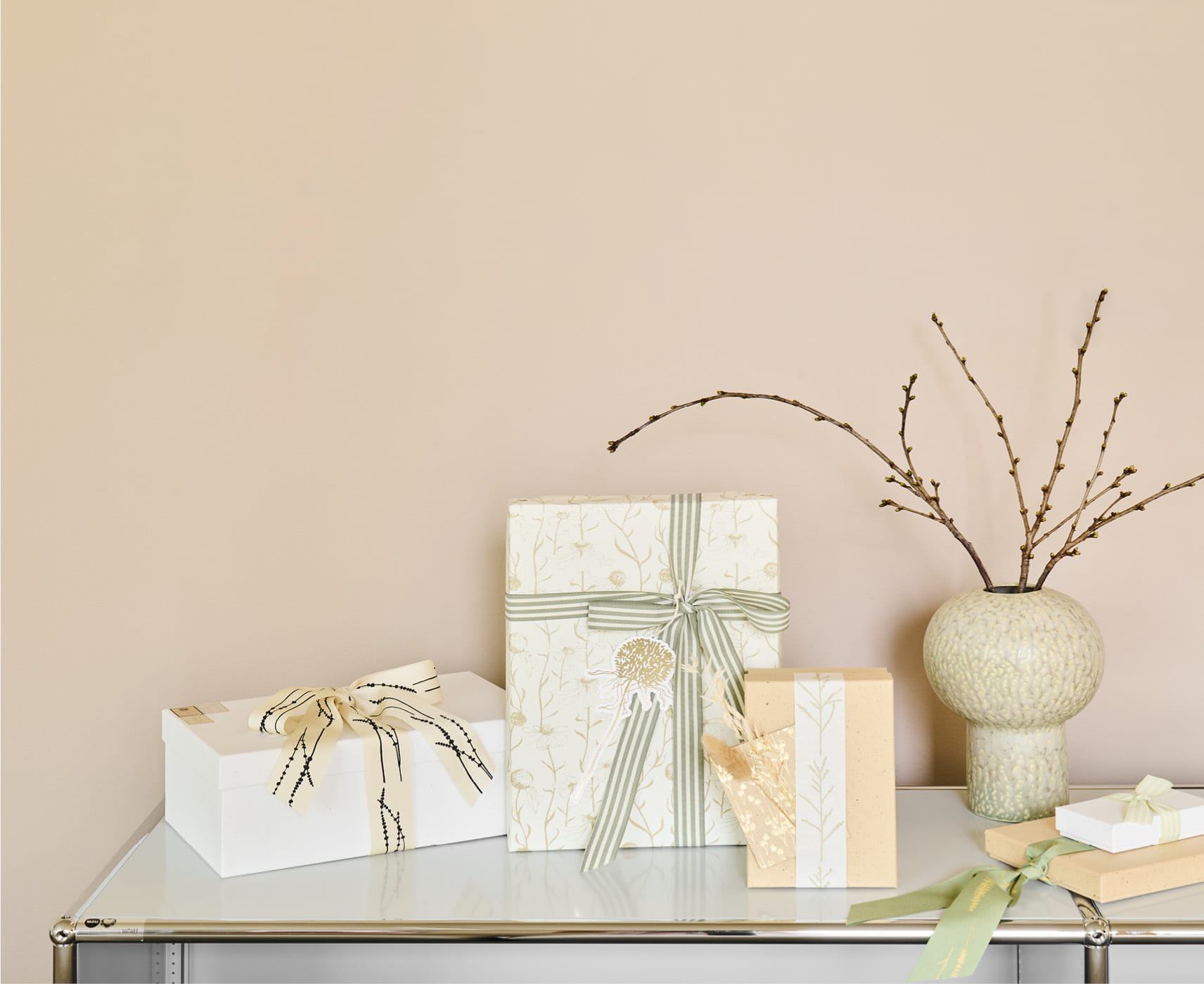 Verpackte Geschenke in Naturtönen und mit Details von Gräsermotiven stehen dekorativ auf einem weißen Regal. 