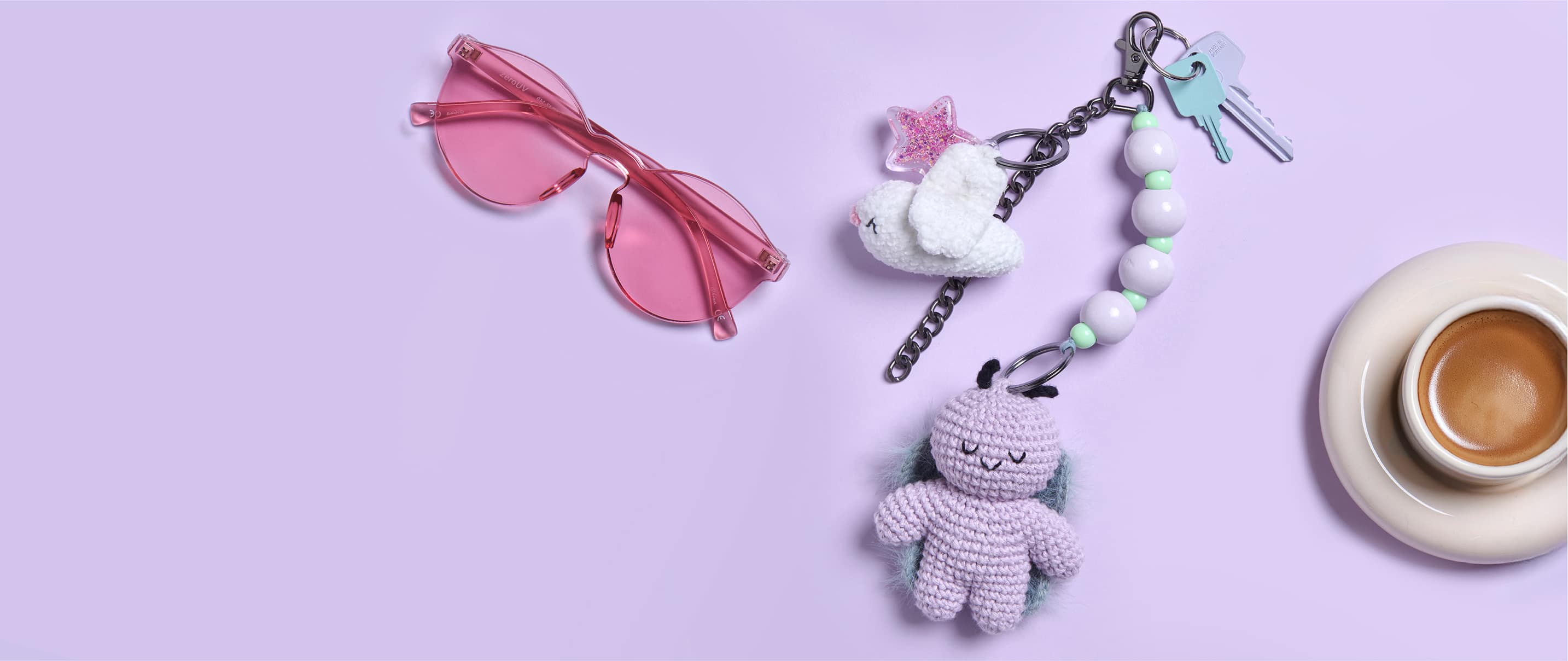 Schlüssel mit gehäkelten Anhängern in Form von einem Vogel und einem Käfer liegen umgeben von einer Sonnenbrille und einer Tasse auf lila Hintergrund.