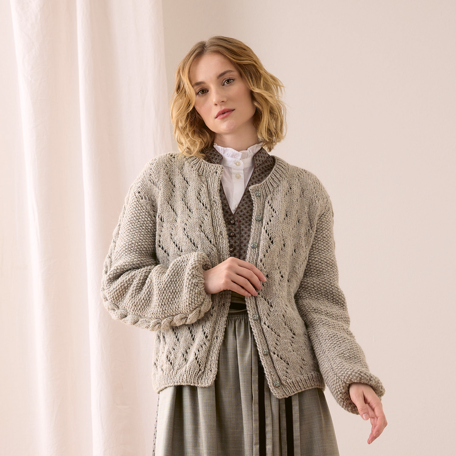 Trachtenjacke aus Soft Alpaca: Elegante, beige Strickjacke mit Zopfmuster, kombiniert mit traditioneller Tracht. Ideal für stilvolle Anlässe.