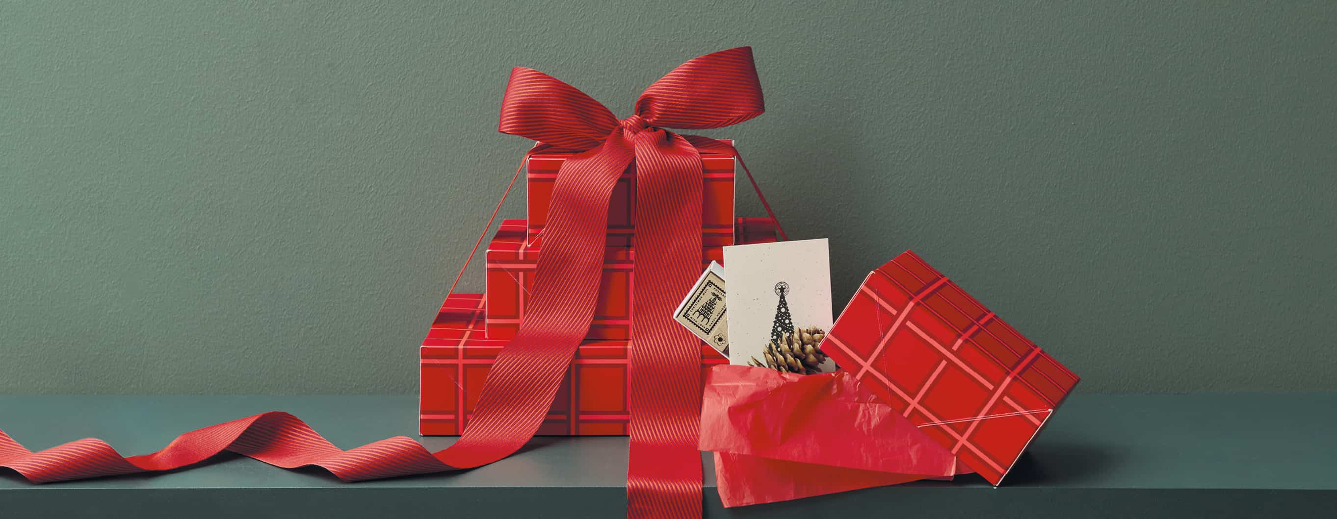 Geschenkboxen in verschiedenen Größen mit einem roten Karomuster liegen gestapelt und mit einer Schleife geschmückt vor einem grünen Hintergrund.