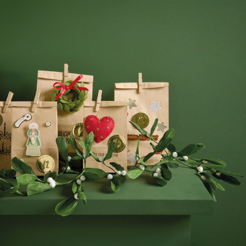 Ein Adventskalender aus Papiertüten ist weihnachtlich dekoriert und steht vor einem dunkelgrünen Hintergrund. 