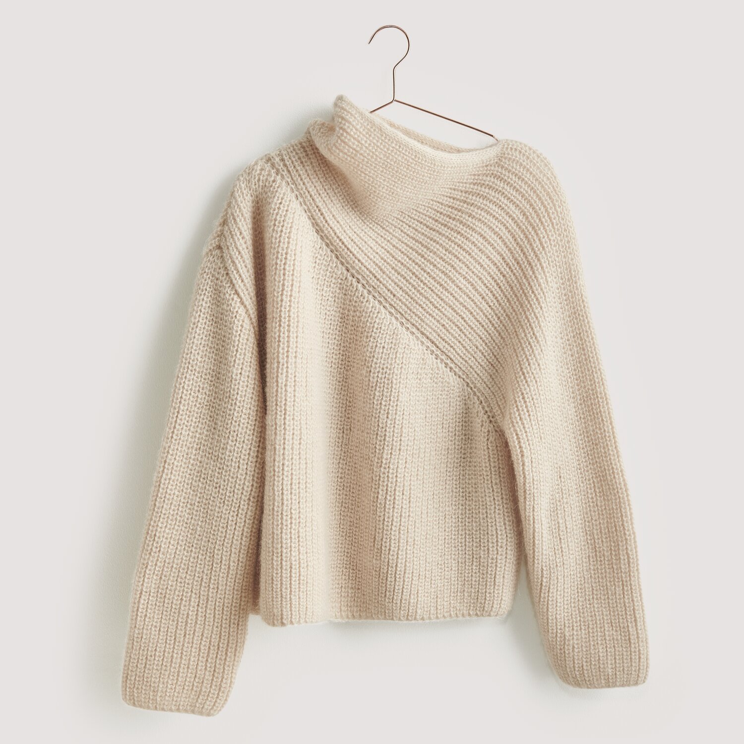 Luxury Knits - Premium Natural Fibres Sprachauswahl deutsch von Rico Design Strickset Pullover Modell 01 aus Luxury Knits: Beigefarbener Pullover mit asymmetrischem Strickmuster, hängend an einem Kleiderbügel.