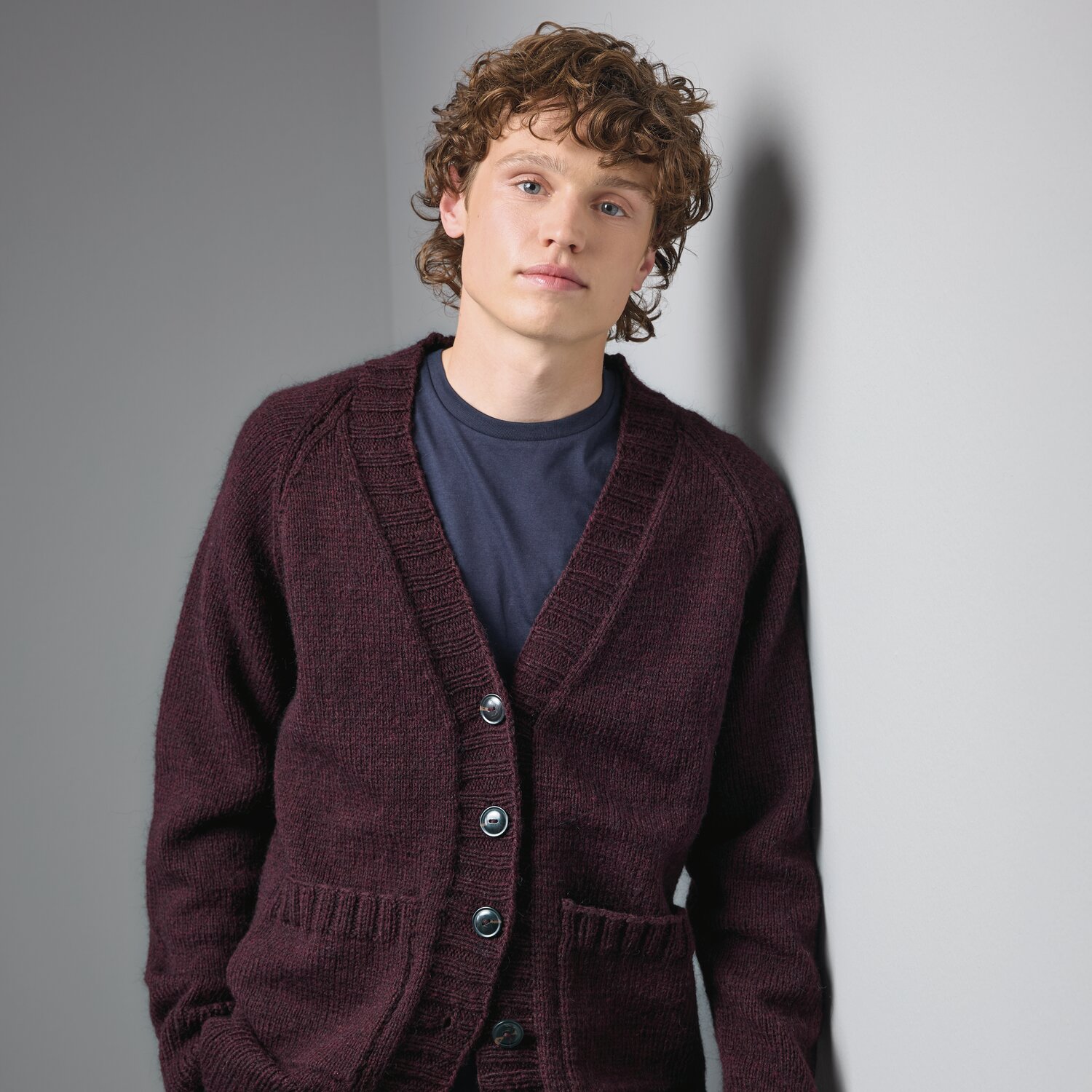 Strickset Jacke Modell 16 aus Made by Me Men No. 3 von Rico Design Strickset Jacke Modell 16 aus Made by Me Men No. 3: Dunkelrote Strickjacke mit Knöpfen, getragen von einem jungen Mann vor grauer Wand.