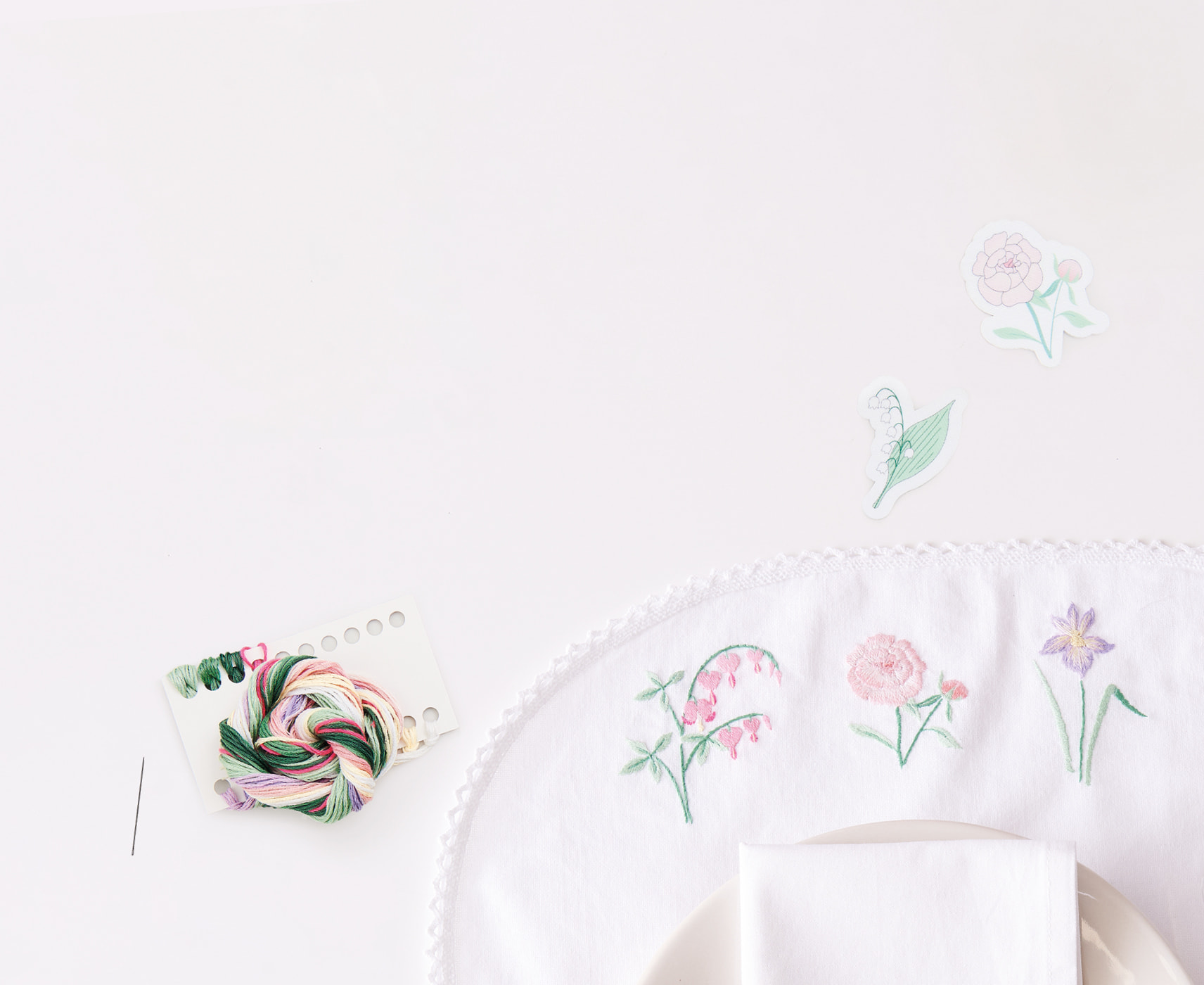 Mit Blumen besticktes Tischset liegt umgeben von floralen Stick-and-Stitch-Vorlagen und Stickzubehör auf weißem Hintergrund.