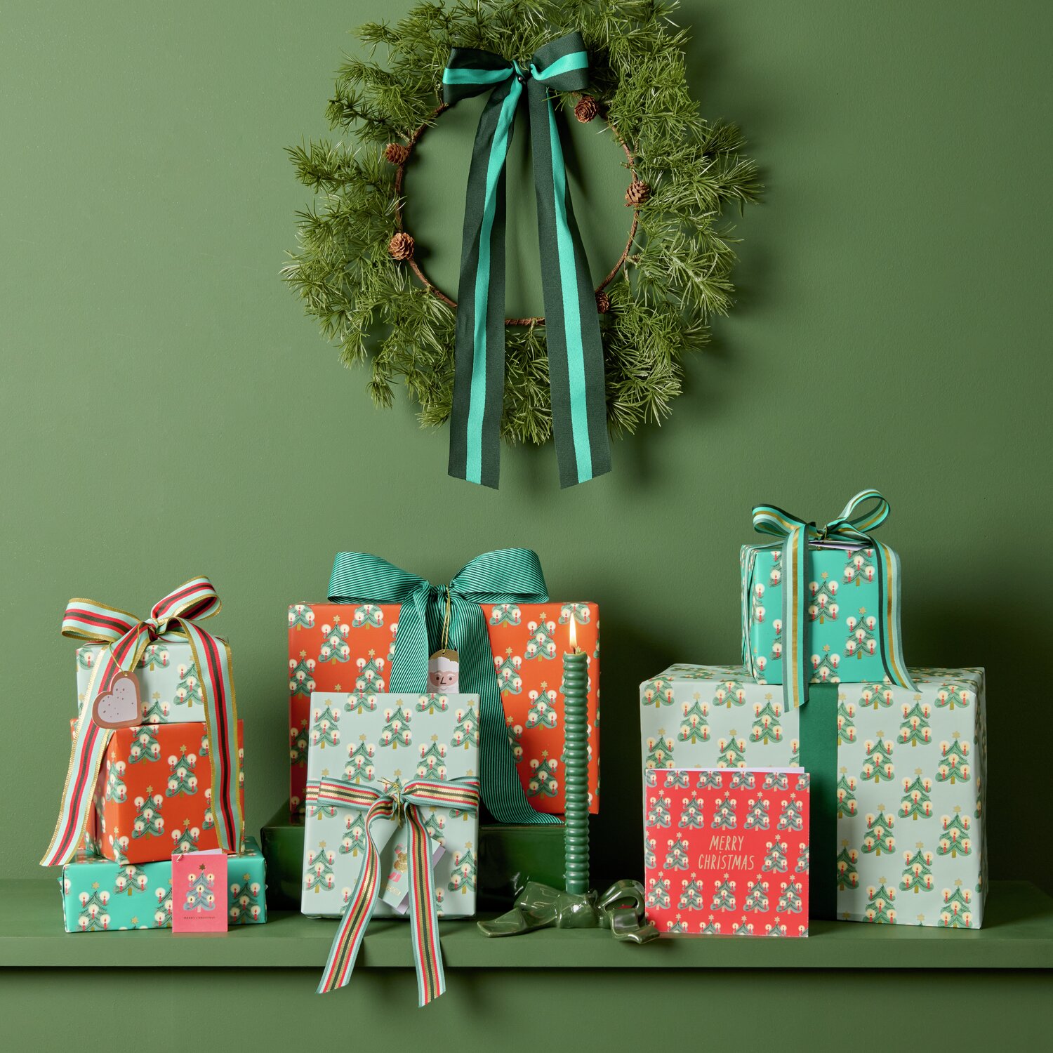 Geschenkverpackungen in verschiedenen Größen, eingewickelt in buntes Paper Poetry Geschenkpapier mit Tannenbaum-Motiven von Rico Design. Farbenfrohe Bänder und eine grüne Kranzdekoration ergänzen das festliche Arrangement.