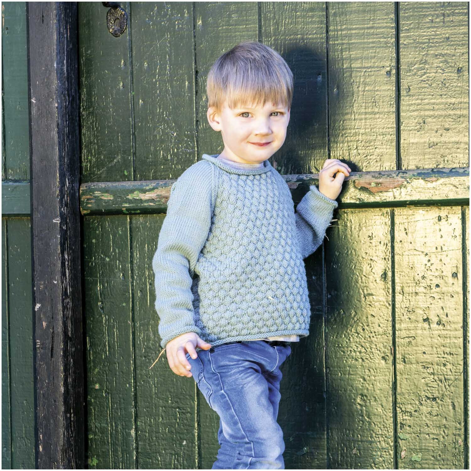 Essentials Merino dk: Kind in gestricktem Pullover vor grüner Holztür, lächelnd und lässig posierend.