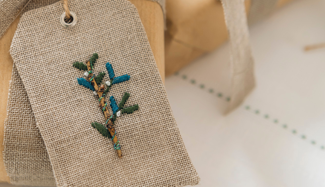 Stickanleitung Geschenkanhänger für Weihnachten: Handgestickter Anhänger auf Leinenstoff mit blau-grünem Zweigmotiv, ideal für festliche Dekorationen.