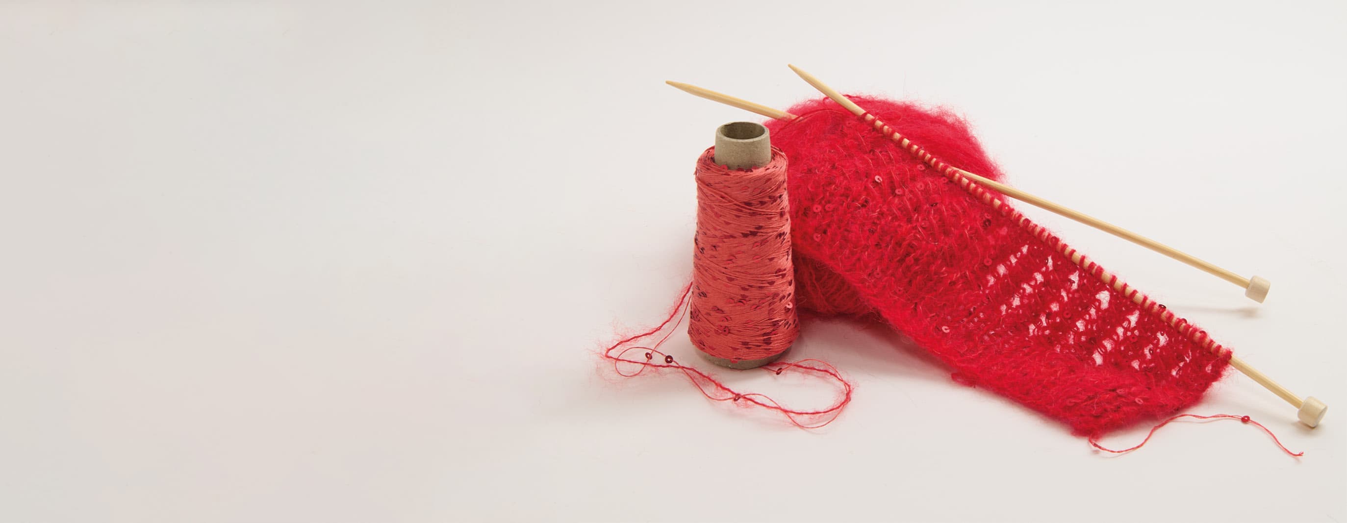 Ein Strickstück auf Stricknadeln steht mit einem roten Paillettengarn und einem roten Garnknäuel vor grauem Hintergrund.
