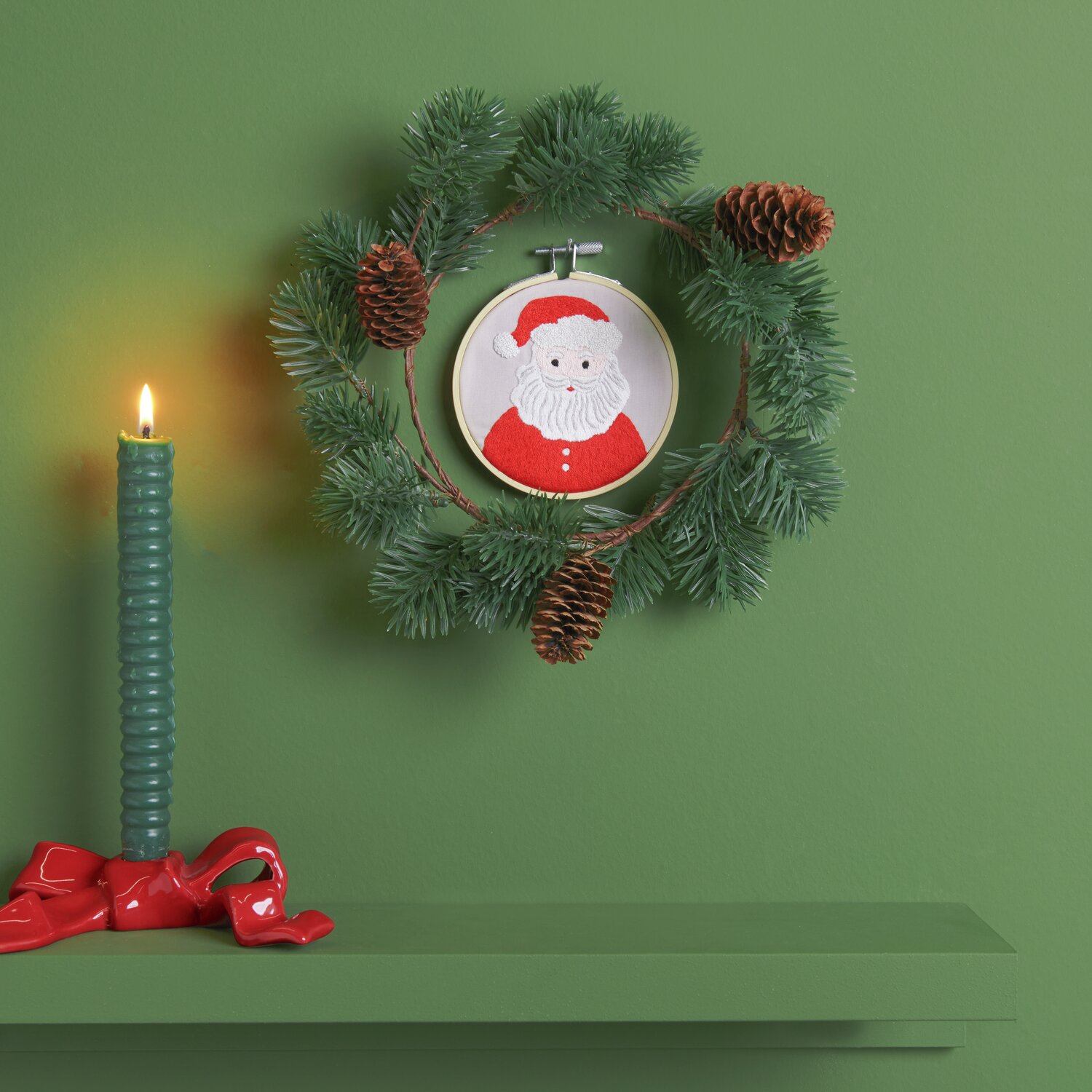 Stickpackung Santa vorgezeichnet Ø10cm von Rico Design Stickpackung Santa vorgezeichnet: Ein weihnachtlicher Kranz mit Tannenzweigen und einem gestickten Santa-Motiv, dekoriert mit Zapfen und einer grünen Kerze.
