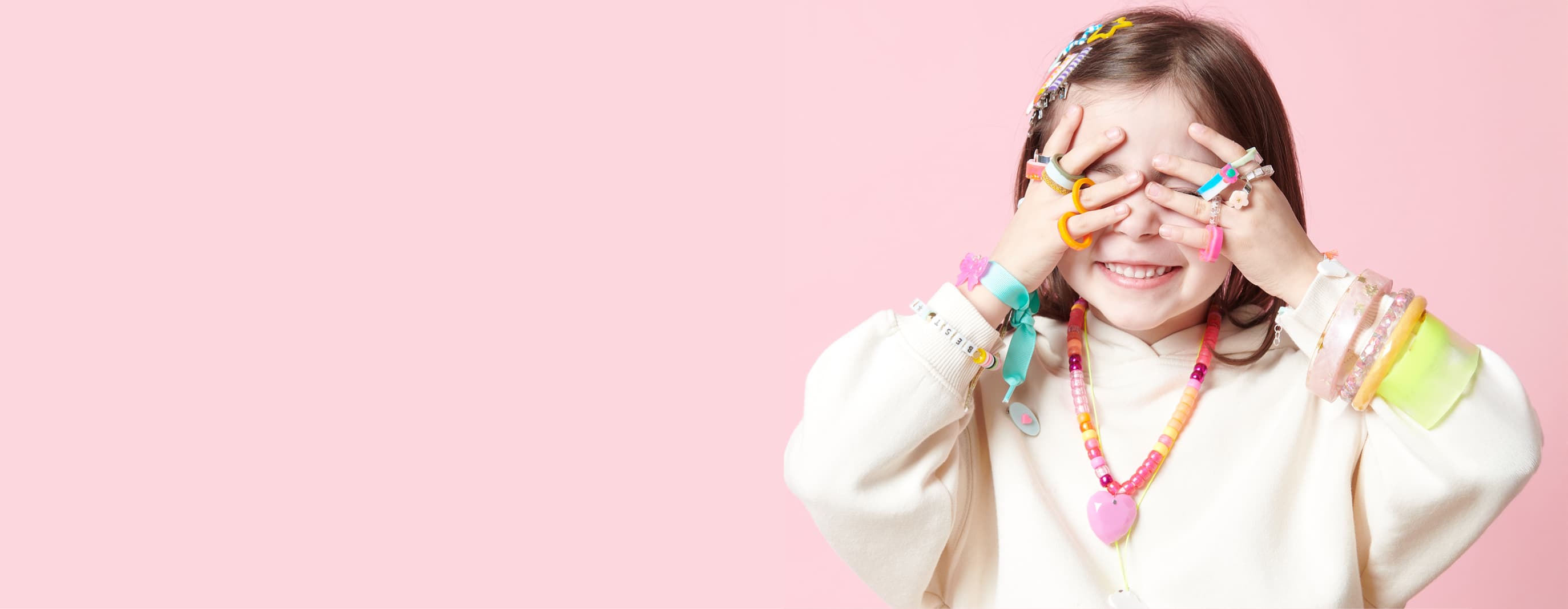 Ein Mädchen steht vor einer pinken Wand und hält ihre Hände vor ihr Gesicht. Ringe, Armbänder und Haarklammern aus UV Resin schmücken ihre Finger, Arme und Haare.  