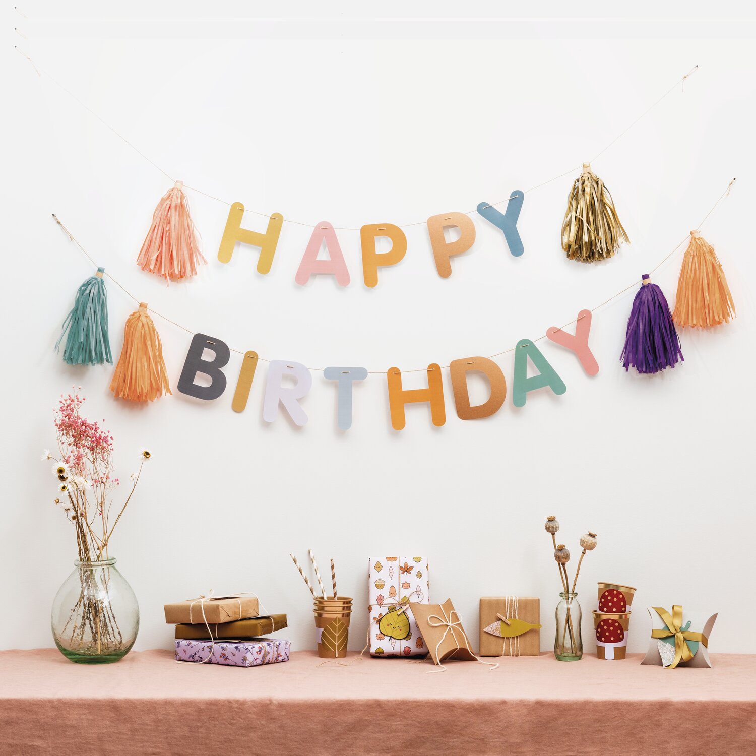 Paper Poetry DIY-Girlande Happy Birthday 3m: Mehrfarbige Buchstaben und Quasten dekorieren eine festliche Wand über einem Tisch mit Geschenken.
