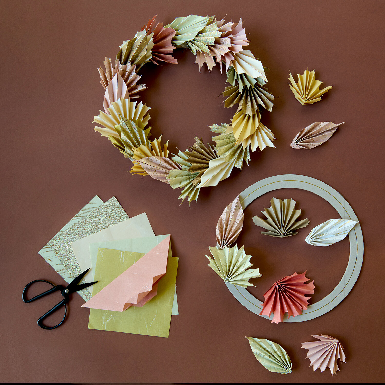 Bastelanleitung Herbstkranz aus Origamipapier, gefaltete Blätter in warmen Pastelltönen mit Schere, Papierbögen und Kranzrohling auf braunem Untergrund