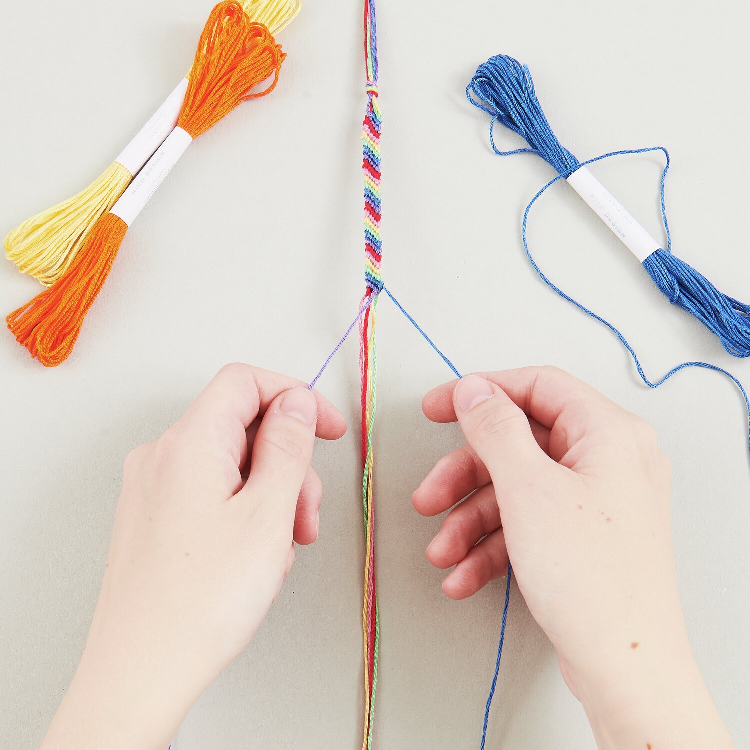 DIY Freundschaftsarmbänder Set Classic Colours: Hände flechten bunte Armbänder mit Garn in Blau, Gelb und Orange.