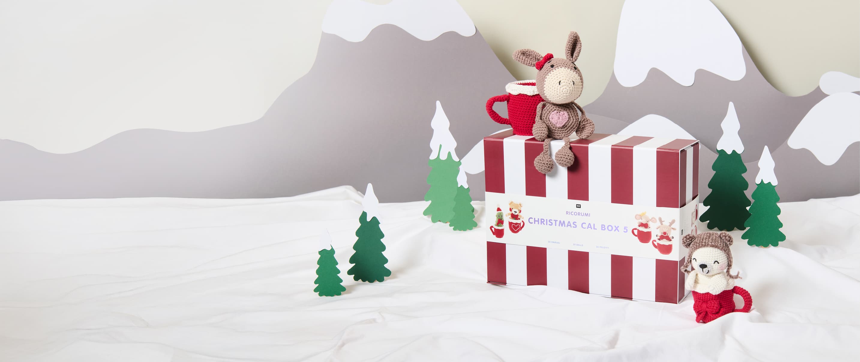 Ricorumi Xmas CAL Box 5 steht in einer Winterlandschaft umgeben von zwei gehäkelten Figuren 