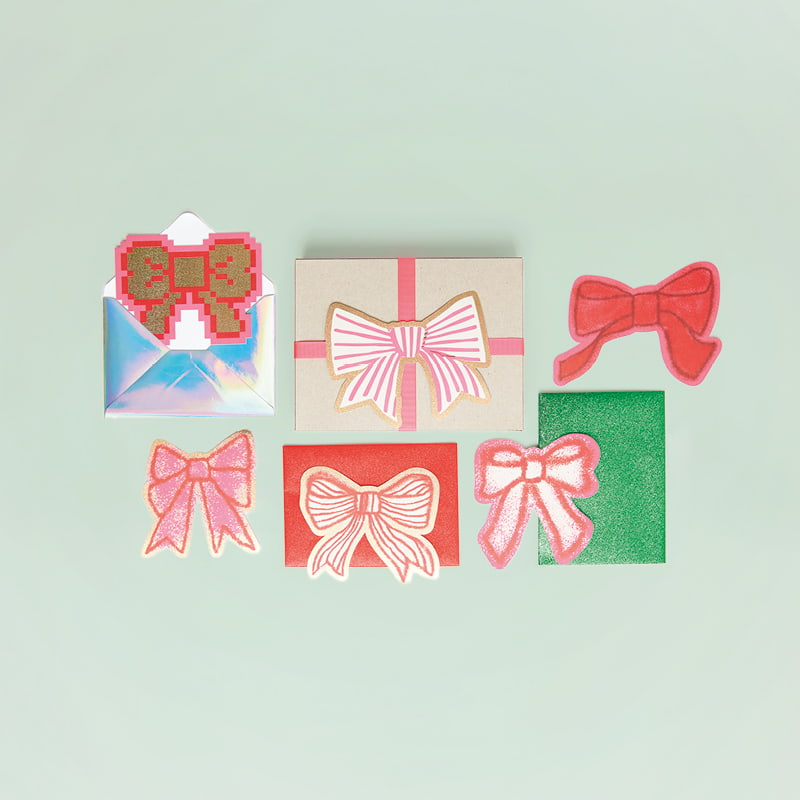 Karten mit Schleifenmotiven in Rot und Rosa liegen mit Geschenkbox und Umschläge auf grünem Hintergrund.